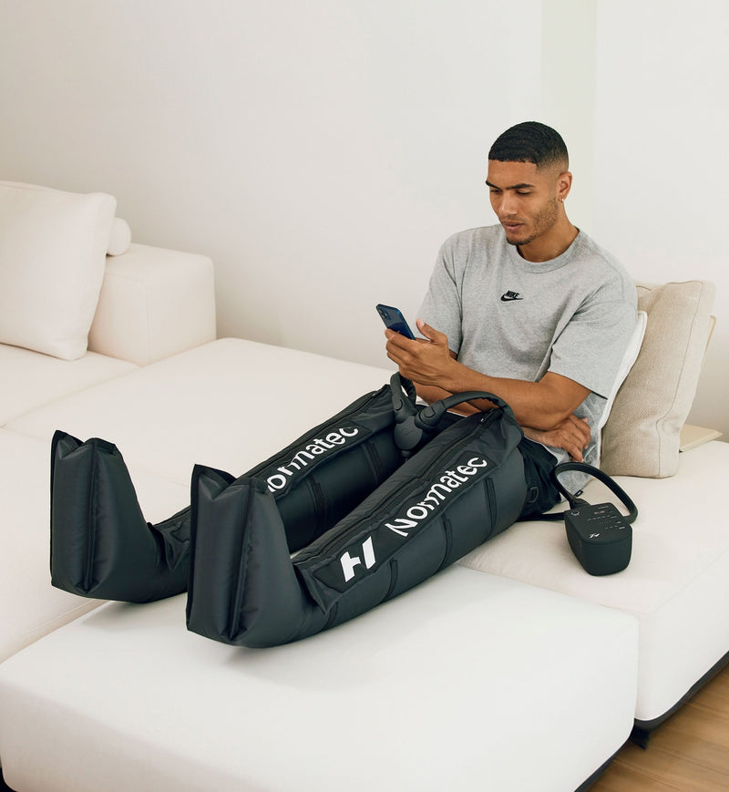 Man sitting on a couch using a phone wearing normatec 3 legs on his legs